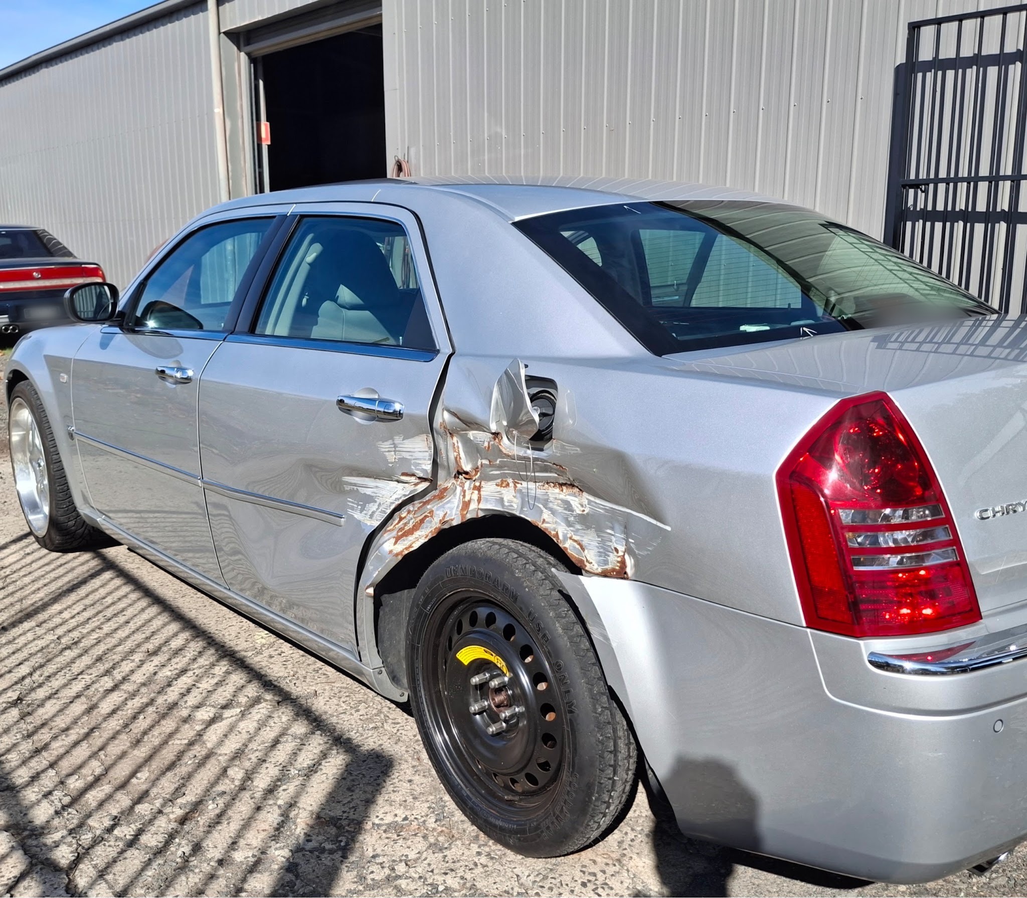 The Chrysler 300C before repair, rear quarter panel torn open behind the wheel arch with a space saver wheel fitted
