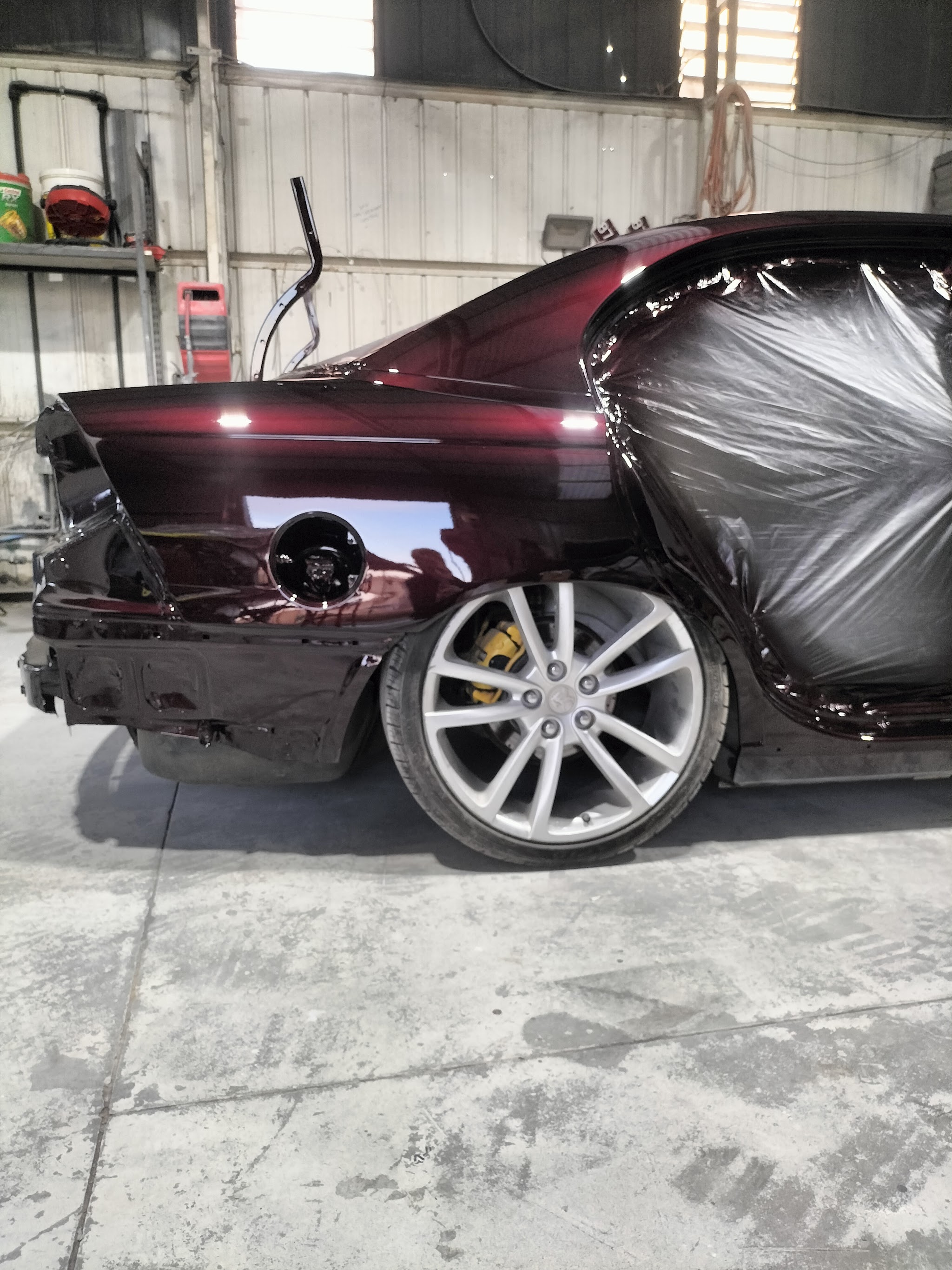 The rear quarter of a black cherry Holden coupe fresh out of paint, still partly sheeted, with yellow brake calipers behind the wheel